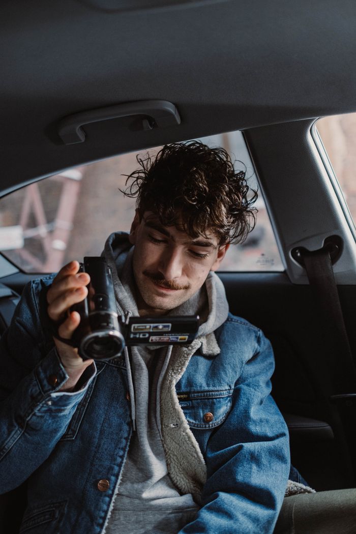 Young man with curly hair filming with a camcorder inside a car.